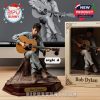 Bob Dylan figure sitting casually with guitar and harmonica brace on decorative rug base next to earthy-toned packaging and 3D render!