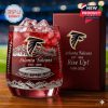 A commemorative whiskey glass with Atlanta Falcons branding, featuring intricate silver mountain and stadium etchings on a red background, placed next to a themed box.