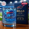 65th anniversary commemorative whiskey glass with Buffalo Bills logo and blue-themed engraving, placed in front of its matching display box on a wooden surface in a stadium setting.