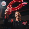 A person raising the Cincinnati Reds 135th Anniversary whiskey glass with ice cubes, celebrating in front of the Reds' neon logo.