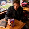 Man in casual jacket sitting near stadium window, holding Clemson Tigers purple whiskey glass beside book on wooden table!