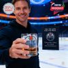 Man in dark hoodie presenting a commemorative Oilers whiskey glass on an indoor hockey rink with promotional display reading