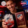 Happy man holding FC Cincinnati whiskey glass and matching box in a bar setting, showcasing team branding and product packaging!
