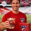 A fan at the stadium smiling and showing off a FC Dallas whiskey glass filled with a red drink, with the team’s shirt and product packaging in view!