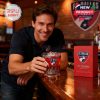 A scene inside a bar where a man happily presents a themed FC Dallas whiskey glass and box under warm lighting!