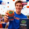 Person holding a Florida Gators whiskey glass outdoors with orange and blue decorations in the background.