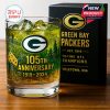 A commemorative Green Bay Packers glass with green drink and ice on a wooden table, next to a black box with gold lettering highlighting the team’s history and logo!