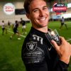 Fan holding Raiders 65th Anniversary glass at practice field in team shirt!```