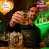 A man toasts with an LAFC whiskey glass in a classy bar, surrounded by bottles and ambient lighting, with a branded gift box on the counter!