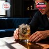 A man in a dark shirt sets down an LAFC-themed whiskey glass filled with ice on a wooden coffee table, surrounded by upscale furnishings!