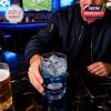 Person holding the Los Angeles Rams whiskey glass at a bar, filled with ice and blue liquid, in front of a football game being shown on TV.