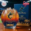 A round whiskey glass shaped like a baseball, printed with the Miami Marlins logo and stitched seams. The glass is paired with a matching collector box featuring team branding and anniversary details!