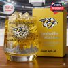 A Nashville Predators collector whiskey glass with gold tones team logo and city imagery stands beside a yellow presentation box in an arena setting!