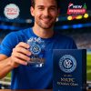 A man in a blue shirt holds a whiskey glass with the NYCFC logo and golden city skyline. A navy blue product box stands beside him on the stadium table!
