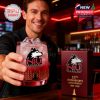 Man at bar holding out the NIU Huskies whiskey glass with anniversary box beside him, smiling in a red-lit space!