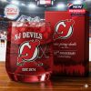 A close-up of a red NJ Devils whiskey glass filled with ice next to a matching gift box, both featuring the team's logo and text highlighting the 52nd anniversary limited edition.