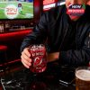 A man enjoys a drink at a sports bar, holding a red NJ Devils whiskey glass while a game plays on a nearby screen, highlighting the product’s festive and game-day appeal.