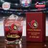A clear red glass featuring Ottawa Senators branding and decorative symbols stands next to a red anniversary box inside a stadium setting!