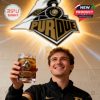 Fan cheering with Purdue Boilermakers whiskey glass under glowing team emblem on interior wall.