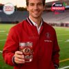 Man holds a San Diego State Aztecs whiskey glass. Red jacket, stadium background. New product offer.!