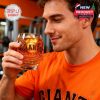 A man in a bright orange Giants shirt sips from a commemorative whiskey glass while standing in a gym, appreciating its craftsmanship and team tribute.