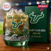 South Florida Bulls whiskey glass with ice and golden beverage on a wooden surface next to a green box displaying the Bulls logo and team message in a softly lit stadium setting!