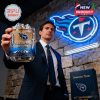 Man in formal wear presenting a Titans glass toward the camera, standing beside a branded gift box and neon Titans logo on a brick wall!