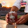 A person presents a Texas State-themed whiskey glass while standing beside a stadium at sunset, emphasizing the commemorative design and school spirit!