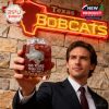 A man in a suit lifts a branded whiskey glass in front of a lit-up Texas State Bobcats sign, symbolizing school pride, tradition, and celebration!