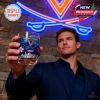 Man in dark blue outfit raising a UVA whiskey glass with blue drink in front of a neon team logo and stone wall background indoors!