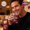 Close-up of a smiling fan enjoying a red beverage from a Wisconsin Badgers whiskey glass at home, surrounded by warm lighting and branded items!