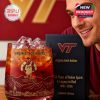 Person smiling while holding a Virginia Tech Hokies whiskey glass with red liquid, with the Hokies anniversary branding clearly visible on the glass.