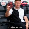 A man in a black and white t-shirt sits in stadium stands, holding up a 148th-anniversary Manchester United glass with ice, featuring Old Trafford design and club crest!