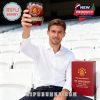 Man in white shirt seated at stadium lifts Manchester United 148th anniversary glass, with gift box beside him on a chair!