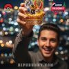 Smiling man holds a Pokémon-themed whiskey glass in focus, featuring Pikachu in a tuxedo and legendary Pokémon under golden anniversary flames, with city bokeh behind!