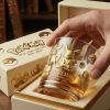 A hand lifting a golden themed Pokemon anniversary glass from a luxury gift box on a wooden table.