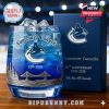 A whiskey glass commemorating the Vancouver Canucks 56th anniversary filled with ice, showcasing team logo, bridge and forest engravings, with packaging displayed behind it.