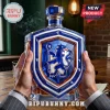 Crystal whiskey decanter shaped like a blue shield with a lion crest held in hands against a blurred bar background.!