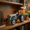 Grey plastic brick tractor model on a wooden bookshelf next to leather-bound books and a brass lamp.!