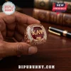 Kappa Alpha Psi fraternity ring with red gemstone, gold lettering, and diamond accents held in a hand over a wooden desk.!