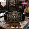 Lynyrd Skynyrd metal belt buckle featuring a skull, wings, and band text on a black leather belt on a wooden desk.!