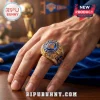 Gold New York Knicks championship ring with blue stone, crystal accents, and engraved team logos on a person's hand.!