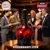 Large ornate red and black glass decanter with a crown stopper on a round wooden table, surrounded by people holding glasses.!