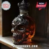 Skull-shaped glass whiskey decanter with an ornate dark metal crown stopper on a wooden shelf surface.!
