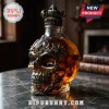 Skull-shaped glass whiskey decanter with an ornate dark metal filigree overlay and crown stopper on a marble table.!