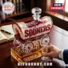 Covered wagon shaped glass decanter labeled Sooners in deep red and cream colors on a wooden desk with a crystal stopper.!