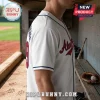 White baseball jersey with red lettering and blue trim worn by a man in a stadium dugout with a leather glove and ball.!