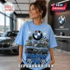 Light blue graphic t-shirt featuring a vintage BMW 76, palm trees, fireworks, and the BMW logo in a dealership showroom.!