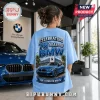 Light blue t-shirt featuring a blue BMW car graphic, 76 years of BMW text, palm trees, and dealership background.!