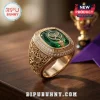 Gold championship ring with a large emerald green stone, tennis racket engraving, and diamond bezel on wood surface.!
